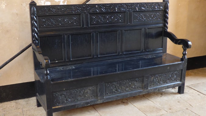 A large wooden bench painted in very dark blue, almost black paint. It is on a stone floor. There are carved patterns on the chest.